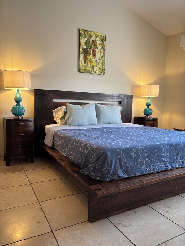 Un dormitorio con una cama de madera, dos mesitas de noche con lámparas, un cuadro en la pared y suelo de baldosas.