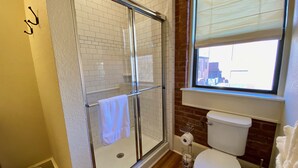 Een badkamer met een douchecabine van glas, een toilet en een raam met uitzicht op gebouwen.