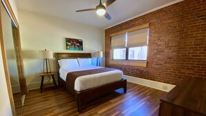 Een slaapkamer met een houten vloer, een bed met witte lakens, een plafondventilator, een raam met jaloezieën en een bakstenen muur.