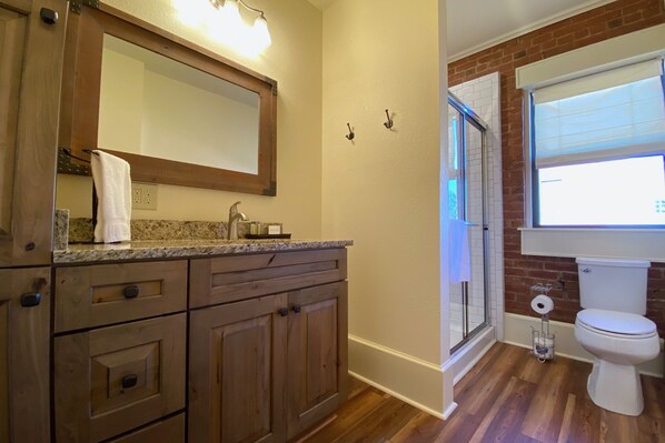 A bathroom with wooden cabinets, a granite countertop, a mirror, a toilet, and a window with blinds.