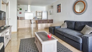 A modern living room with a grey sofa, a coffee table, and a TV on a stand. There is a kitchen area in the background with a dining table and chairs. - Appartement Gimelli – Welkeys (Toulon)