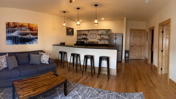 A modern living room with a dark grey sofa, a wooden coffee table, and a kitchen area with a bar and stools. - Main Street Retreats 105 - Brand New Apt. 105 (Waynesville)