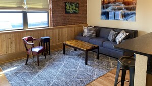 A cozy living room with a wooden floor, a gray sofa with patterned cushions, a wooden coffee table, a leather chair, and a large window with blinds. - Main Street Retreats 105 - Brand New Apt. 105 (Waynesville)