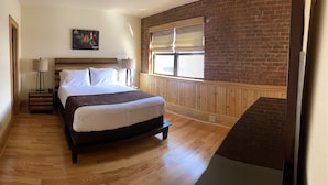 A bedroom with a wooden bed, a nightstand, a lamp, a window with blinds, and a brick wall. - Main Street Retreats 105 - Brand New Apt. 105 (Waynesville)