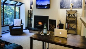 Una sala de estar con chimenea, un televisor montado en la pared, una computadora portátil sobre una mesa y una estantería.
