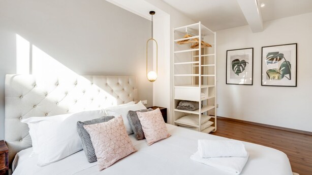 A bedroom with a tufted headboard, a white bed with patterned pillows, a white shelving unit, and framed artwork on the wall.