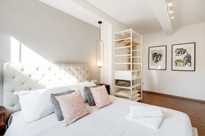 Un dormitorio con cabecera capitonada, cama blanca con almohadas estampadas, estantería blanca y cuadros en la pared.
