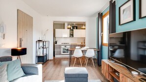A modern living room with a sofa, ottoman, and a TV stand. There is a dining area with a table and chairs, and a kitchen in the background.