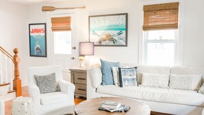 Een woonkamer met een witte bank, een ronde salontafel en een witte fauteuil. Er hangen lijstjes met foto's aan de muur, een lamp en een raam met jaloezieën.