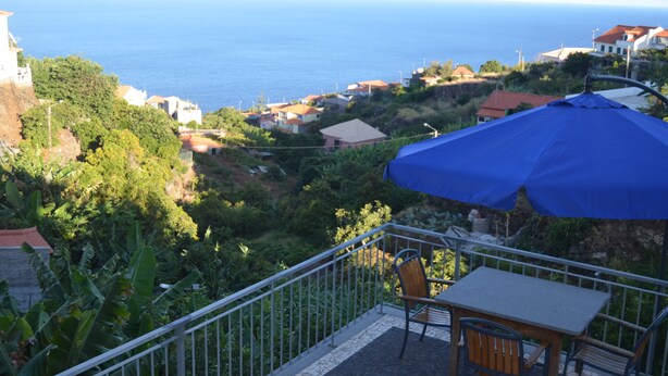 A balcony with a table and chairs, overlooking a coastal area with houses and greenery. - Descobrir Madeira com Carro Grátis (Estrada Padre Alfredo Vieira Freitas)