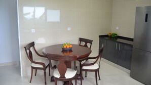 A dining area with a round wooden table surrounded by four chairs, a bowl of fruit on the table, and a modern kitchen with a stainless steel refrigerator. - Descobrir Madeira com Carro Grátis (Estrada Padre Alfredo Vieira Freitas)