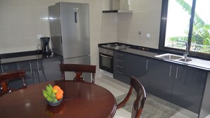 A modern kitchen with a stainless steel refrigerator, a gas stove, and a sink. There is a dining table with chairs and a bowl of fruit. - Descobrir Madeira com Carro Grátis (Estrada Padre Alfredo Vieira Freitas)