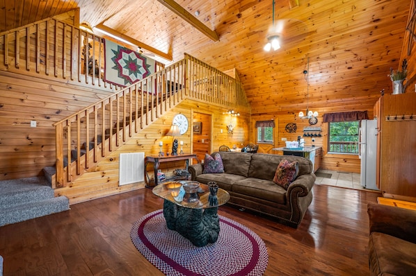 A wooden cabin interior with a staircase, a living room with a sofa, a coffee table, and a kitchen area.