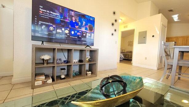 Una sala de estar con un televisor de pantalla plana montado en la pared, un mueble de madera debajo y una mesa de centro de vidrio con un cuenco decorativo.