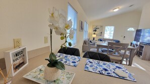 Una habitación bien iluminada con una mesa de comedor, sillas, una orquídea en maceta y un cartel enmarcado en la pared.