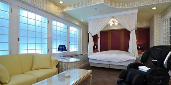 Una habitación con sofá amarillo, una mesa de centro de vidrio, cama con dosel, sillón de masaje y una mesita de noche con lámpara.