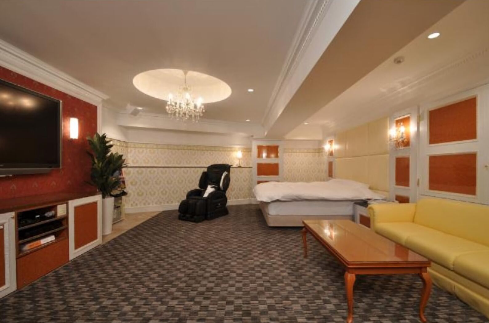 A hotel room with a large flat-screen TV, a massage chair, a bed, a yellow sofa, and a wooden coffee table.
