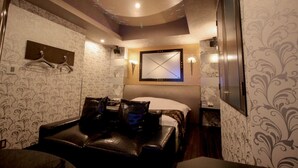 Una habitación de hotel con cama, zona de estar con sofá de cuero, televisor montado en la pared y lámpara en el techo.