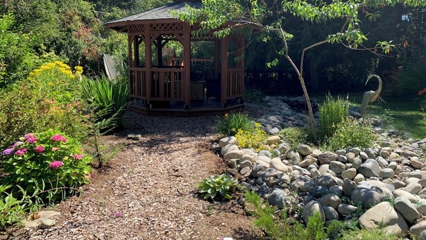 Un pabellón de madera rodeado por un jardín con diversas plantas y un camino de grava.