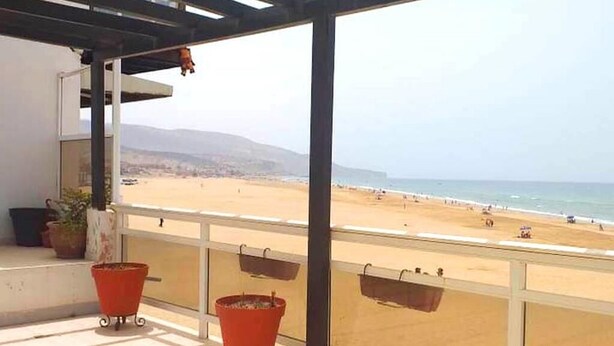 Un balcón con macetas, con vistas a una playa donde hay gente y un cielo despejado.