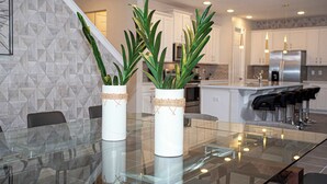 Una cocina moderna con armarios blancos, una mesa de comedor de vidrio y dos plantas en macetas de mimbre.