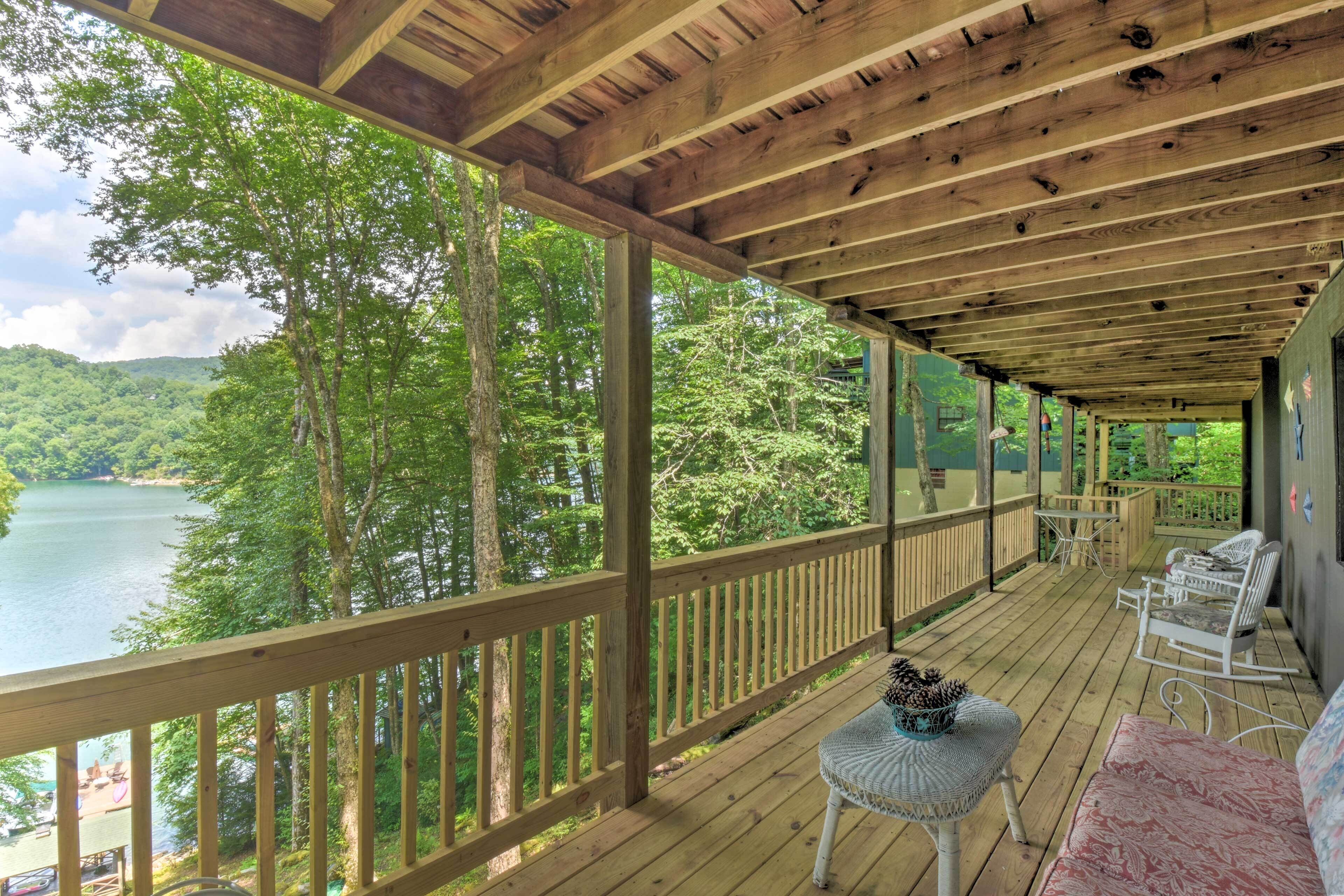 A wooden deck with a view of a lake and forest, white wicker furniture, and a covered area with a wooden ceiling.