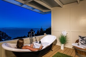 A bathtub with a view of the sea, a bottle, and a glass on the edge.