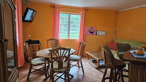 Ein Essbereich mit Holztisch und Stühlen, einem an der Wand befestigten Fernseher und einem Fenster mit Vorhängen.