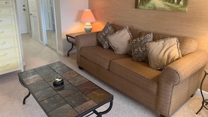 A living room with a brown sofa, patterned cushions, a small table with a lamp, a fish wall decoration, and a painting of a forest. - 10-202 (Banner Elk)
