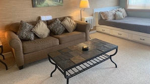 A cozy room with a brown sofa, a black coffee table, a painting of a forest, a lamp, and a bed with patterned pillows. - 10-202 (Banner Elk)