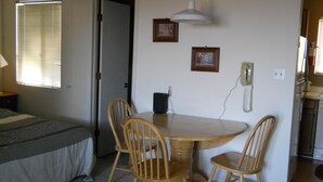 Ein kleiner, kompakter Raum mit einem Bett, einem Esstisch mit vier Stühlen, einem Telefon und zwei gerahmten Bildern an der Wand.