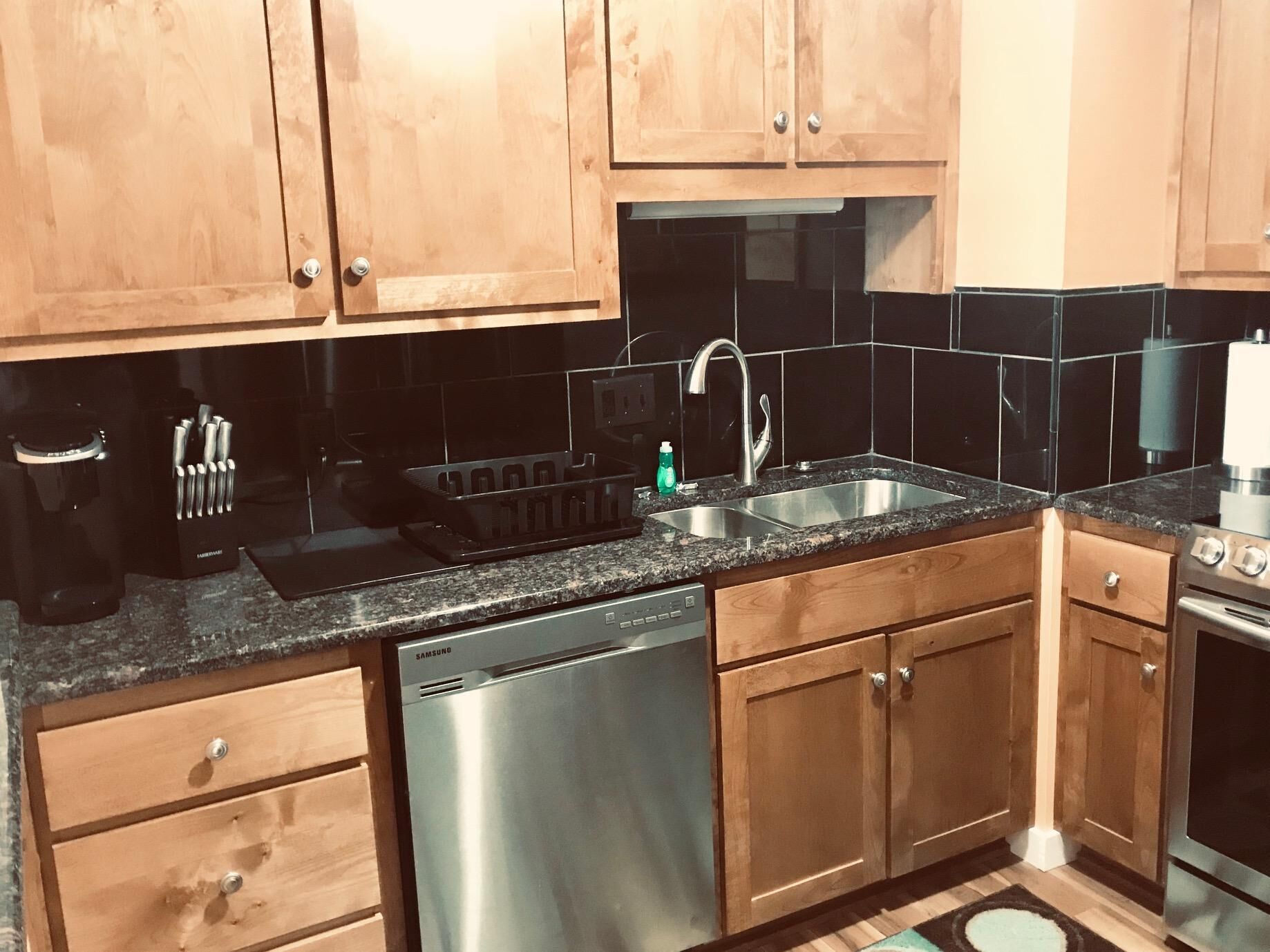 A kitchen with wooden cabinets, a stainless steel dishwasher, a sink with a faucet, and a countertop with a coffee maker and utensils.