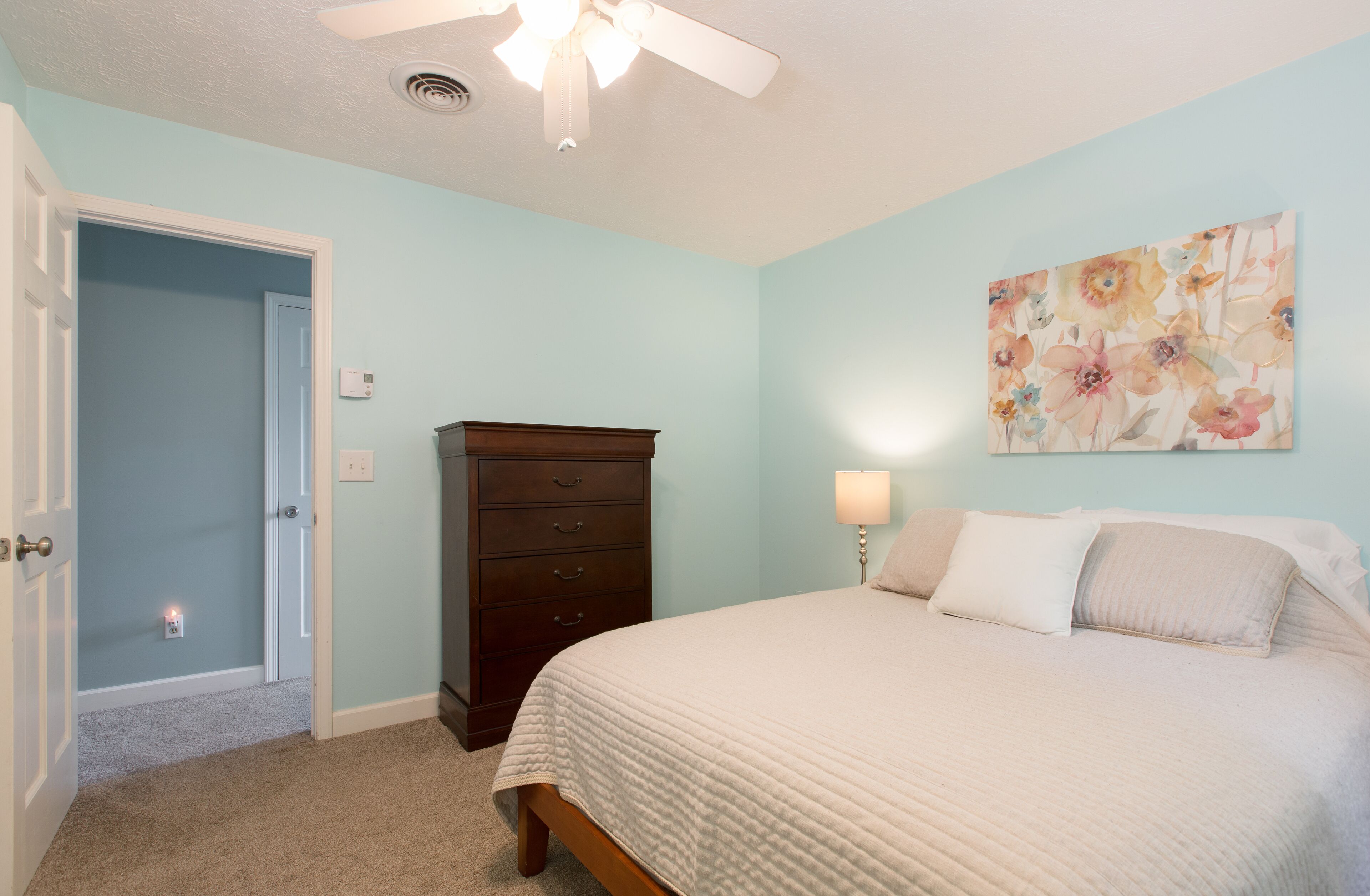 Ein Schlafzimmer mit Bett, Kommode, Deckenventilator und einem floralen Gemälde an der Wand.