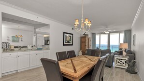A spacious dining area with a wooden table and chairs, a kitchen with white cabinets, and a view of the ocean through large windows. - Pearl 704 Sugar White Sand and Emerald Waters. Spectacular view (Navarre)