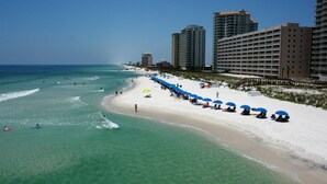 A beach with clear turquoise water, a sandy shore, and several high-rise buildings in the background. - Pearl 704 Sugar White Sand and Emerald Waters. Spectacular view (Navarre)