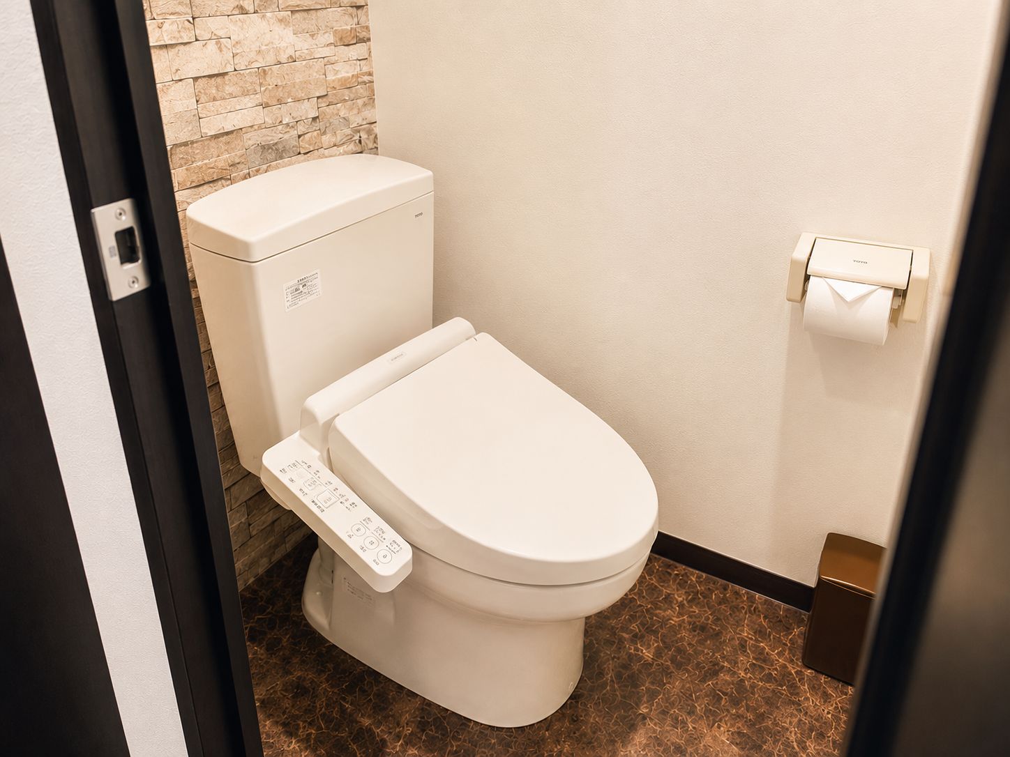 A modern toilet with a control panel, a toilet paper holder, and a stone wall in the background.