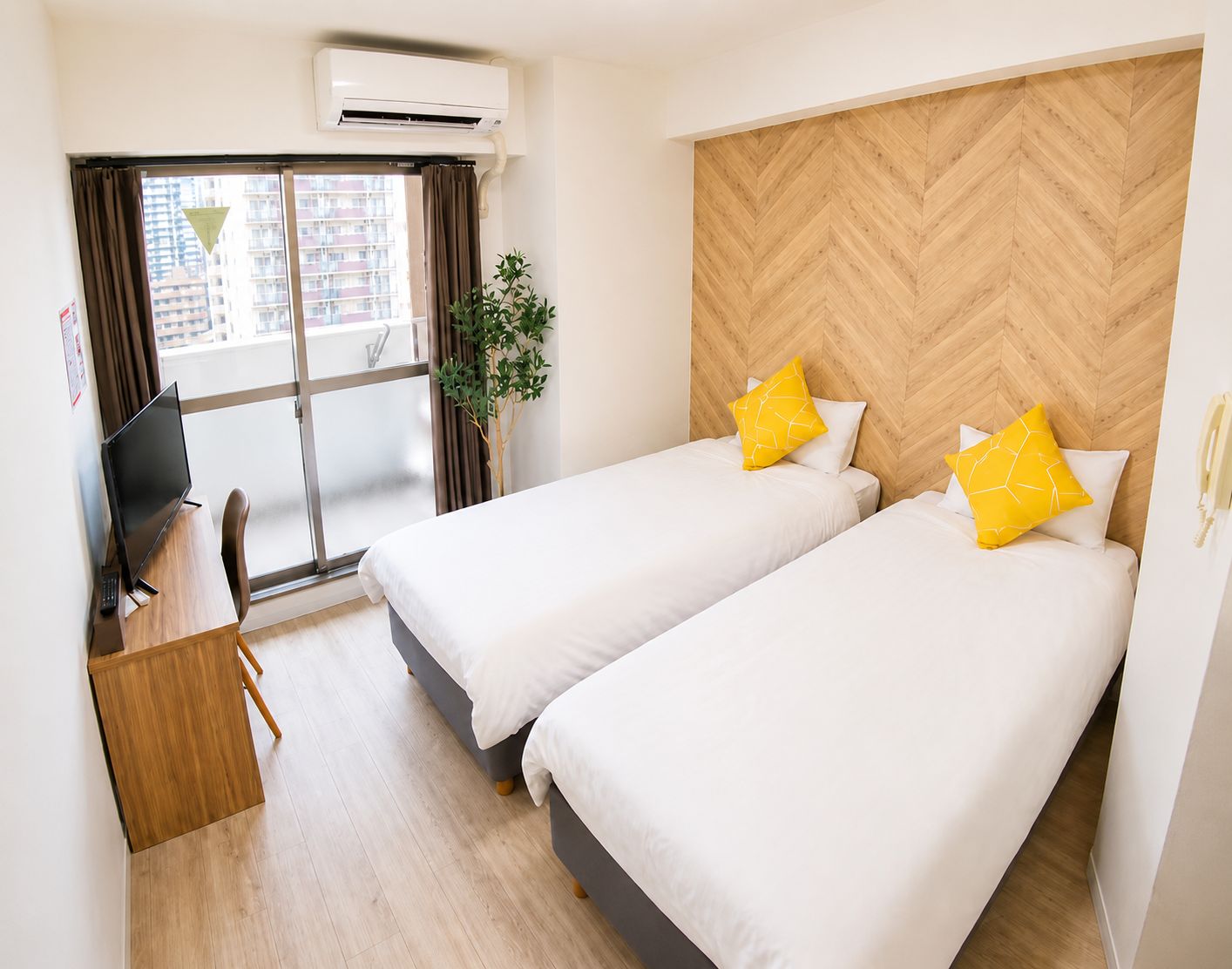 A hotel room with two beds, a desk, a chair, an air conditioning unit, and a window with curtains.