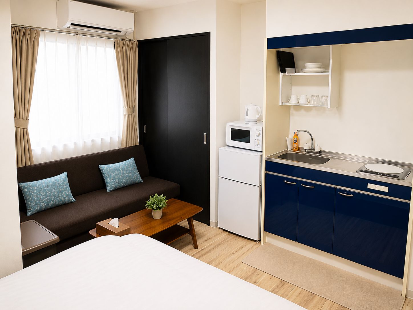 Quarto de hotel pequeno e funcional, com sofá, uma mesinha com planta, cozinha compacta equipada com micro-ondas e pia.