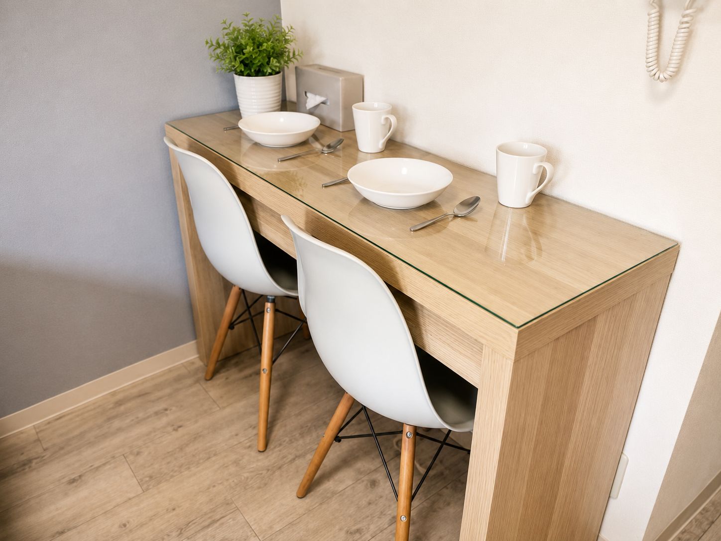 A dining area with a glass-top table, two white chairs, a potted plant, and a telephone on the wall.