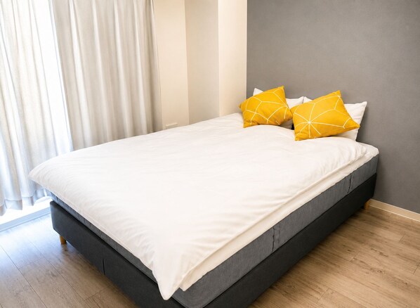 A bed with white bedding and two yellow pillows, a grey wall, and a wooden floor.