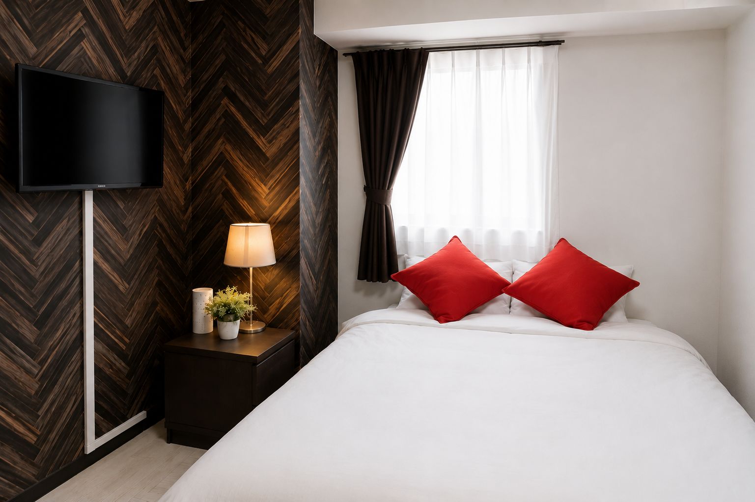 A hotel room with a large bed, a flat-screen TV mounted on the wall, a bedside table with a lamp, and a window with curtains.