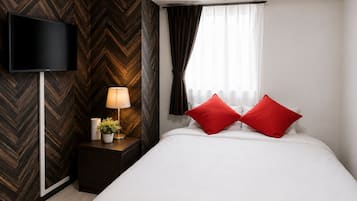 A hotel room with a large bed, a flat-screen TV mounted on the wall, a bedside table with a lamp, and a window with curtains.