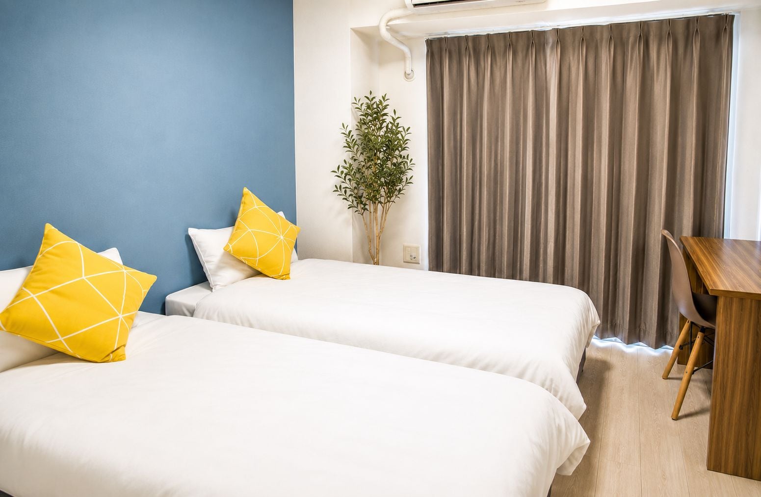A hotel room with two beds, a desk, a plant, and a window with curtains.