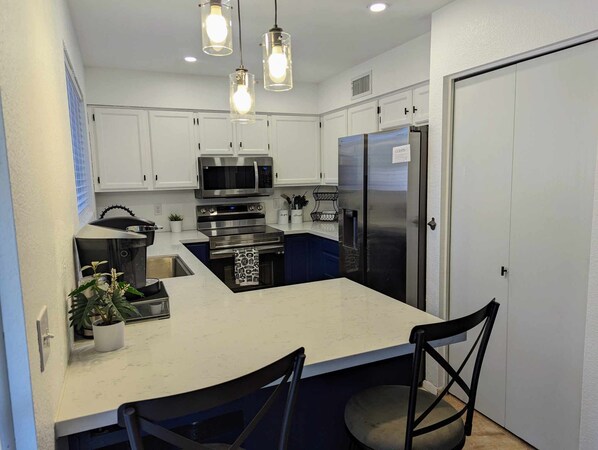 A compact kitchen with white cabinets, a stainless steel refrigerator, a microwave, and a stove. There are two pendant lights above the island, and a potted plant on the counter.