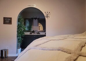 A bedroom with a large bed, a powder room sign, a mirror, and a potted plant.