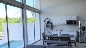 Ein modernes Wohnzimmer mit einer grauen Eckcouch, einem Essbereich mit Tisch und Stühlen, einer Küche mit weißen Schränken und Geräten sowie einem großen Fenster mit Blick auf Grünflächen.