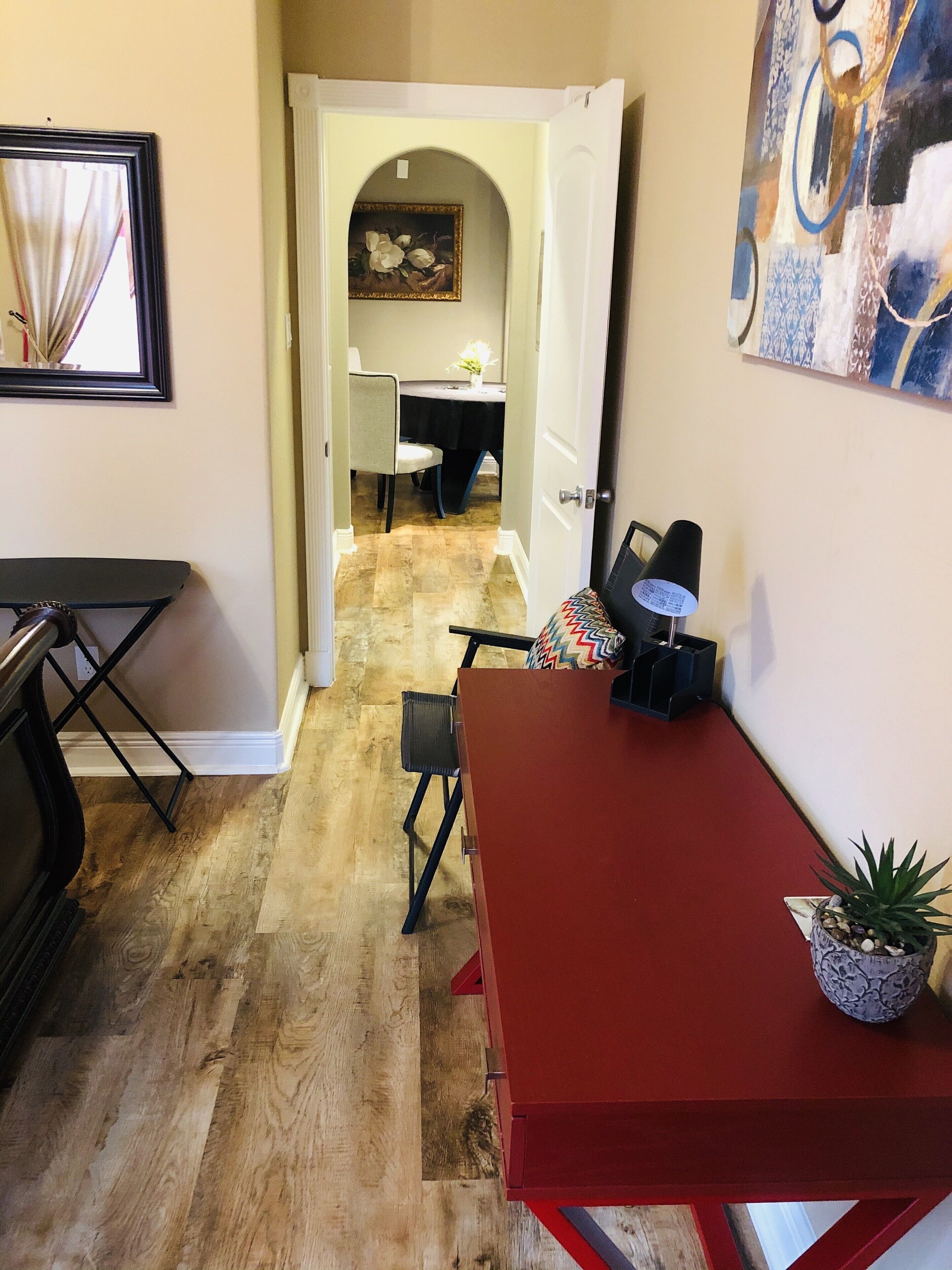A dining area with a red table, chairs, and a potted plant. There is a doorway leading to another room with a table and chairs.