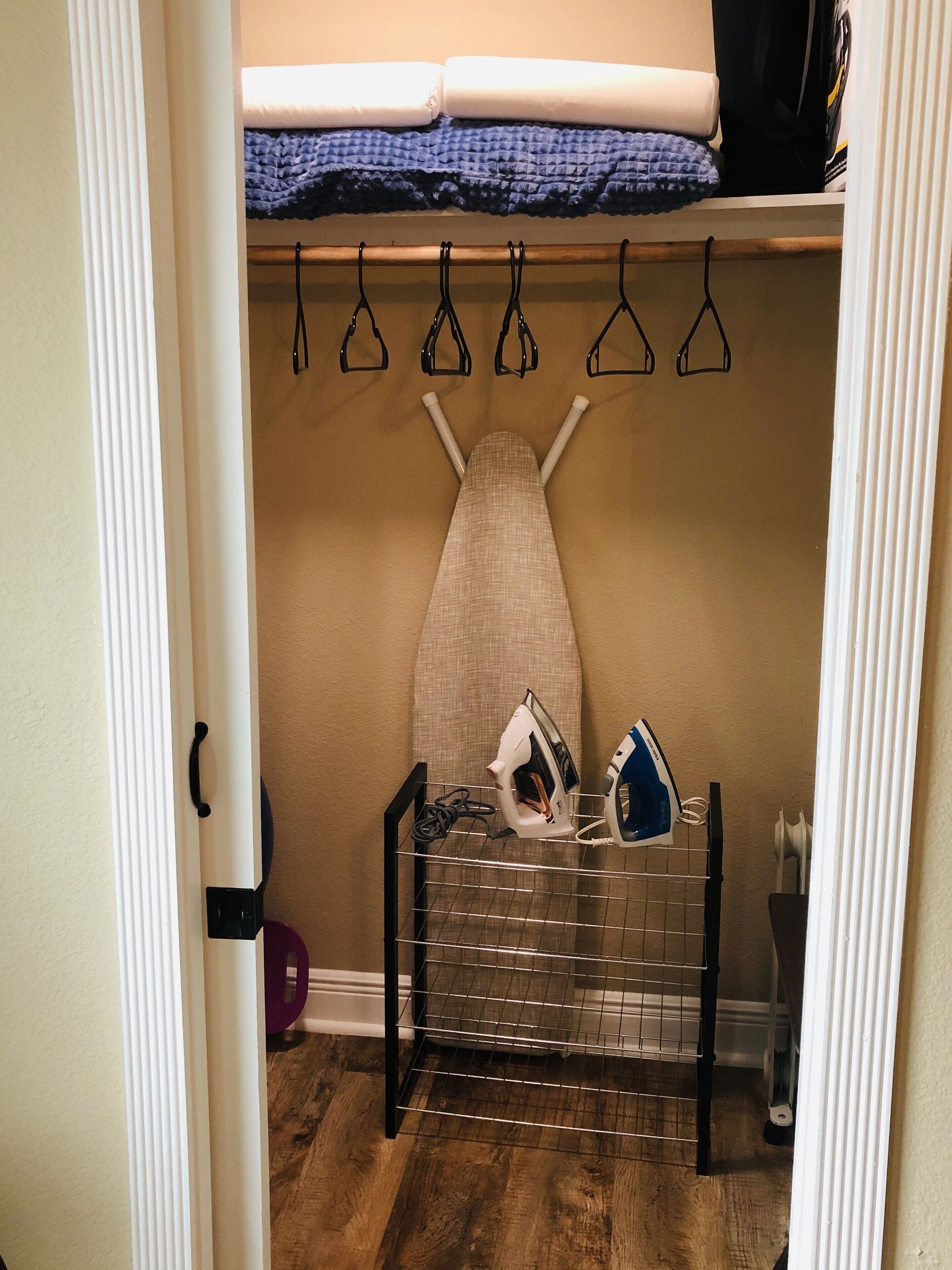 A closet with hanging clothes, an ironing board, and a shelf with folded clothes.