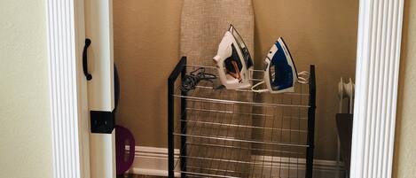 A closet with hanging clothes, an ironing board, and a shelf with folded clothes.