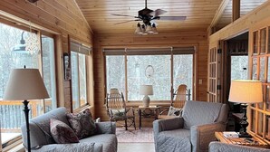 A cozy cabin interior with wooden walls, a ceiling fan, and a mounted deer head. There are two sofas, a red chair, and a patterned rug.